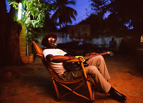 Ali Farka Touré sentado en una silla fumando y con una guitarra en las manos, en Niafunké