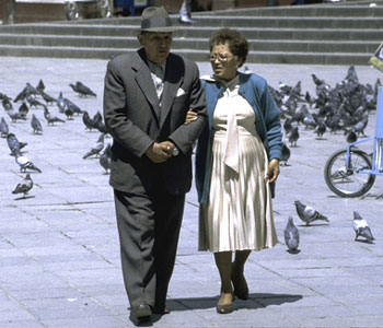 Una pareja de ancianos pasean agarrados por una plaza llena de palomas